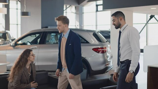 Full shot of polite Middle Eastern car salesman welcoming young Caucasian couple into showroom, inviting to sit on couch, shaking hands with husband, and giving them copies of loan agreement to read