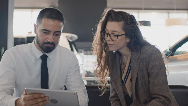 Medium shot of young Caucasian female buyer sitting on couch in car dealership, Middle Eastern male sales manager showing available colours on tablet, girl liking it, smiling, nodding and pointing
