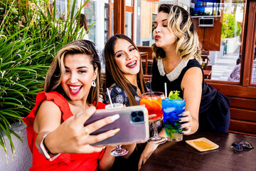 Female friends taking selfies with a mobile phone while enjoying drinking cocktails together.