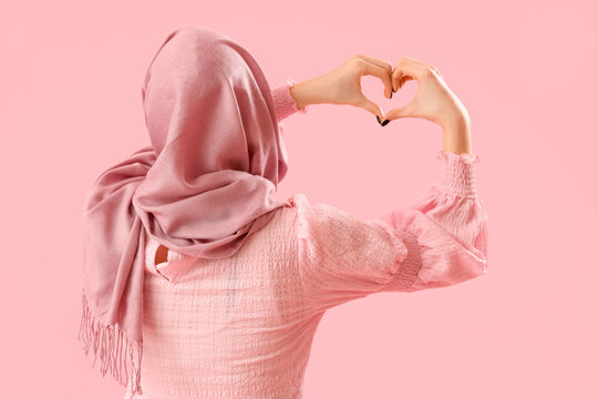 Beautiful young Muslim woman making heart with her hands on pink background, back view