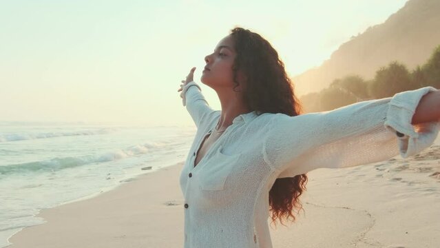 Young attractive ethnic Indian millennial woman posing at sunset spreading arms to sides and inhaling sea breeze dressed in white casual clothes stands on ocean with sandy beach. Love for travel