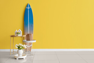 Tables with flowers, books and surfboard near color wall in room