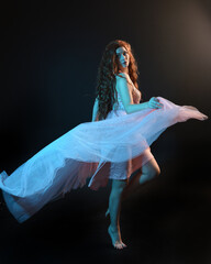 Full length  portrait of beautiful brunette woman dancer, wearing ethereal gown, dancing with flowing fabric.  isolated on dark studio background with cinematic colourful lighting.