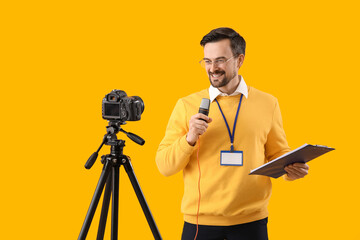 Male journalist with microphone and clipboard recording video on yellow background