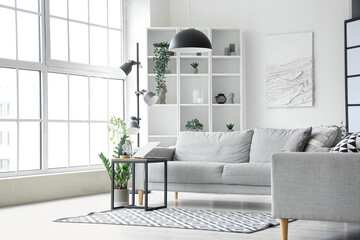 Interior of light living room with cozy grey sofa and modern laptop on coffee table