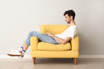 Fototapeta premium Young man working with laptop in armchair near light wall