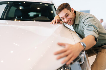 Portrait of happy smiling customer male hugging, stroking car hood after purchased in dealership,...