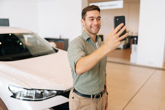 Portrait of happy handsome buyer male client doing selfie picture on smartphone at dealership after bought new car. Smiling young man choosing new vehicle in showroom and making photo using phone.
