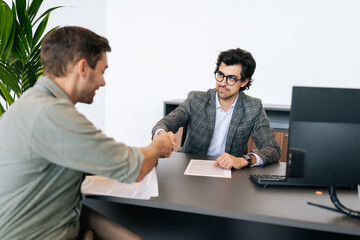 Professional male real estate broker wearing business suit present and advise young man client on decision to sign insurance contracts. Happy guy customer signing purchase agreement and shaking hands.