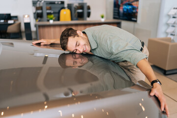 Side view of happy excited customer male in casual clothes hugging stroking auto hood with closed eyes that purchased in dealership. Satisfied young man embracing hood of new car in showroom.