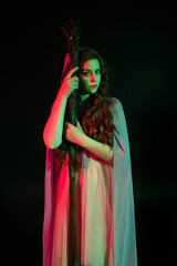 Fototapeta premium portrait of beautiful brunette woman wearing a gown with purple fantasy cloak holding a wooden wizard staff, isolated on dark studio background with cinematic colourful lighting. 