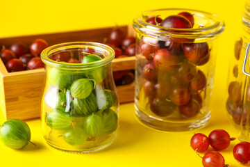 Jars and wooden box with fresh gooseberries on yellow background