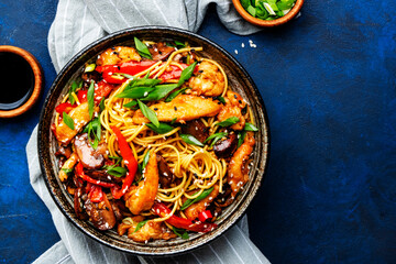 Stir fry egg noodles with chicken, paprika, mushrooms, chives and sesame seeds in ceramic bowl. Asian cuisine dish. Blue stone table background, top view