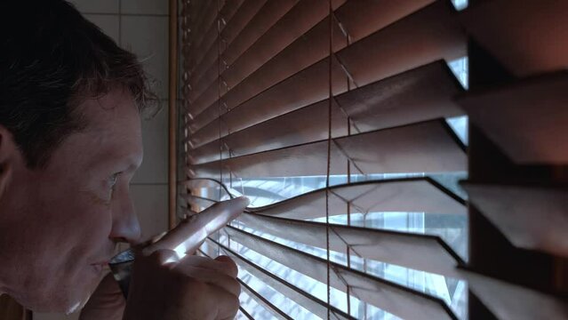 Man Opens The Blinds To Look Out The Window And Drinks Coffee