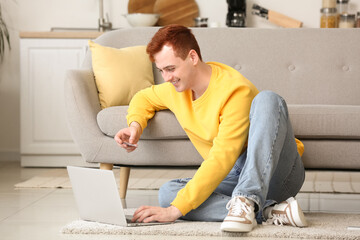 Handsome young man with credit card and modern laptop sitting on floor near sofa while shopping...