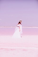 Woman in pink salt lake. She in a white dress and hat enjoys the scenic view of a pink salt lake as she walks along the white, salty shore, creating a lasting memory.