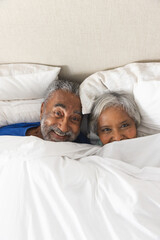 Portrait of happy senior biracial couple lying in bed under bedclothes at home, copy space