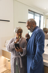 Happy senior biracial couple using smartphone and brushing teeth in bathroom at home