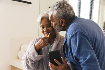 Happy senior biracial couple using smartphone and brushing teeth in bathroom at home