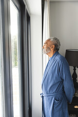 Thoughtful senior biracial man wearing bathrobe and looking through window at home