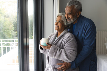 Happy senior biracial couple wearing bathrobes and looking through window with mug of coffee at home