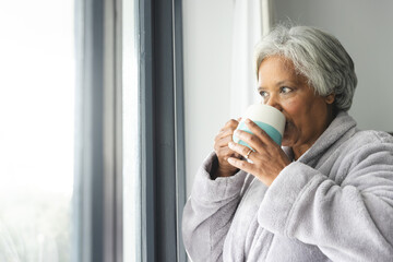 Thoughtful senior biracial woman wearing bathrobe and drinking coffee at window at home - Powered by Adobe
