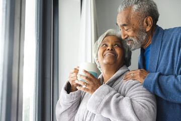 Happy senior biracial couple wearing bathrobes and embracing with mug of coffee by window at home