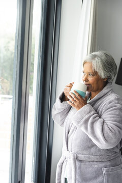 Thoughtful senior biracial woman wearing bathrobe and drinking coffee at window at home
