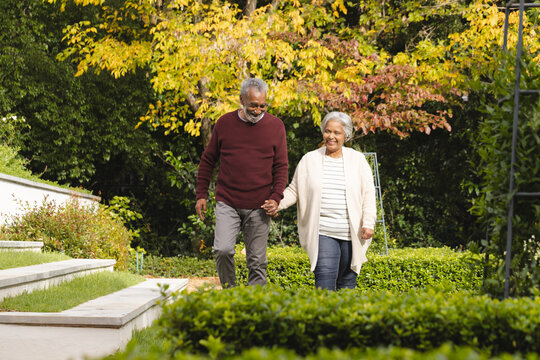 Happy senior biracial couple holding hands and walking in garden at home