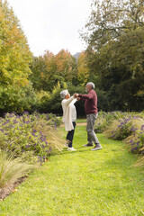 Happy senior biracial couple dancing with flowers around them in garden at home, copy space