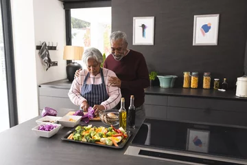 Fotobehang Happy senior biracial couple embracing and preparing vegetables in kitchen at home © WavebreakMediaMicro