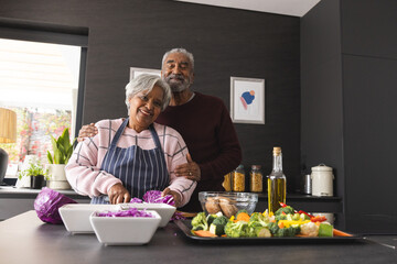 Portrait of happy senior biracial couple preparing vegetables and embracing in kitchen at home
