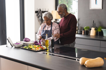 Happy senior biracial couple preparing vegetables and embracing in kitchen at home