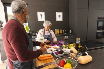 Fotobehang Happy senior biracial couple wearing aprons preparing vegetables and using tablet in kitchen at home © WavebreakMediaMicro