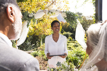 Happy biracial female marriage officiant with tablet and senior couple at wedding in sunny garden