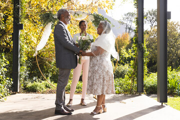 Happy biracial female marriage officiant and senior couple during wedding in sunny garden