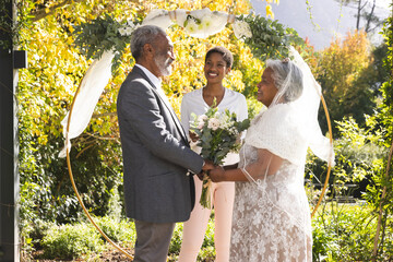 Happy biracial female marriage officiant and senior couple during wedding in sunny garden