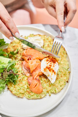 Woman cuts Zucchini pancakes, with avocado, cheese cream, salmon and egg . Healthy breakfast, protein. Restaurant dish. Breakfast in cafe on a sunny morning, on the summer veranda