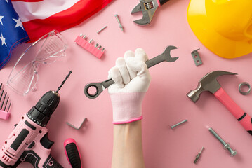 Female worker empowerment symbol. First person overhead view of woman's gloved hand, tightly gripping wrench atop USA flag. Surrounding tools include drill, hammer, screws, safety gear. Pink backdrop