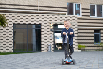 A blonde boy in a white T-shirt, black jacket and black pants riding a scooter. 