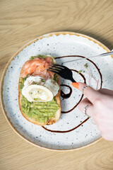 Toast with salmon and a poached egg on an avocado spread top view. Female hands with a fork