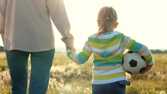 young girl happily walking hand hand with mother soccer field during sunset. girl holding football symbolizing love for sport. both have smiles faces, representing joy fulfillment feel family. scene