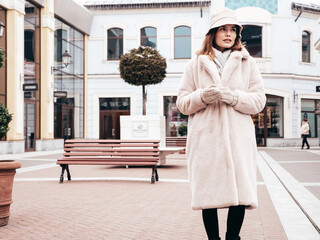 Young beautiful smiling lady wearing trendy white faux fur coat. Stylish woman posing in the street in winter. Cheerful and happy model in panama. Cold autumn weather. In shopping mall