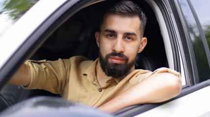 Portrait of a young man with a beard driving his white car. A handsome guy with a beige shirt looks through the car window directly into the camera.