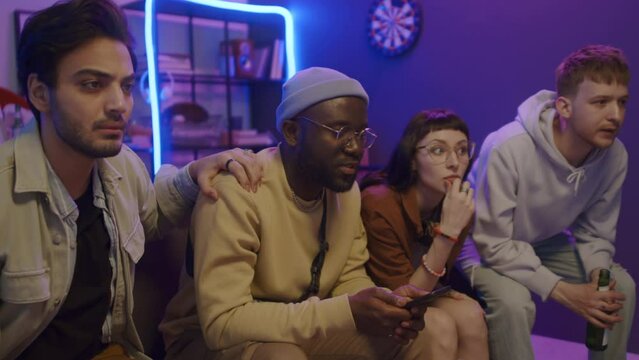 Medium shot of multicultural young male and female students sitting in living room in dorm, anxiously watching sport match on TV, cheering, then reacting with disappointment at missed goal