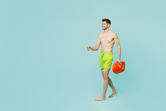 Full body side view young lifeguard man wear green shorts swimsuit relax near hotel pool hold lifebuoy walk go look aside isolated on plain blue background. Summer vacation sea rest sun tan concept.
