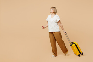 Full body traveler elderly woman 50s wear casual clothes hold bag suitcase isolated on plain beige background. Tourist travel abroad in free spare time rest getaway. Air flight trip journey concept.