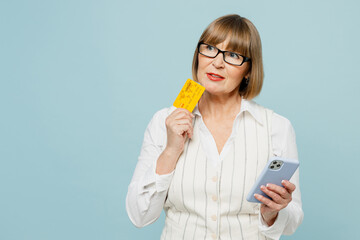 Employee business woman 50s wears white classic suit glasses formal clothes use mobile cell phone hold credit bank card shopping online isolated on plain blue background. Achievement career concept.