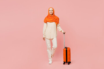 Full body traveler arabian woman wear abaya hijab hold bag suitcase isolated on plain pink background Tourist travel abroad in free time rest getaway Air flight trip, uae middle eastern islam concept