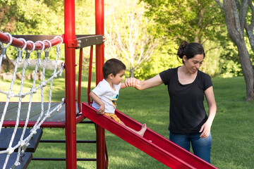mother in her thirties holds her two year old son by the hand as he goes down the slide